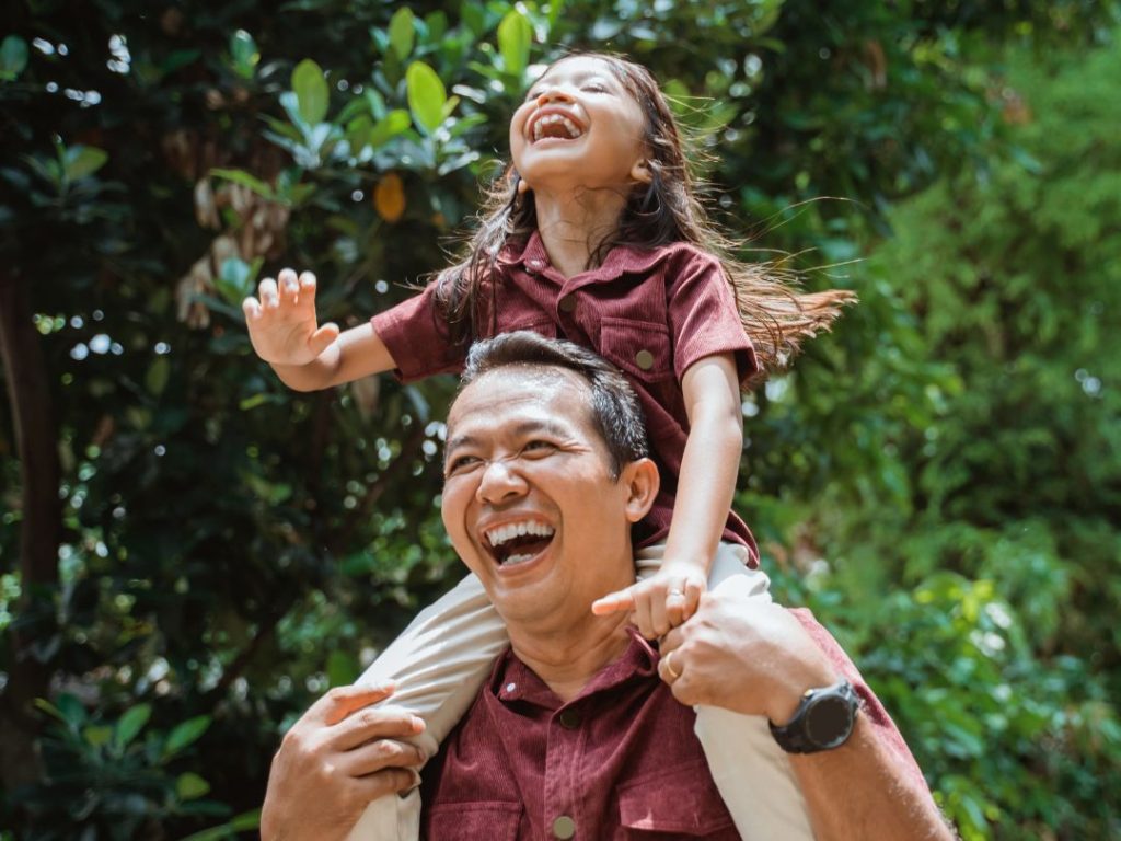 A man carries a laughing young girl on his shoulders outdoors, both wearing matching maroon shirts, surrounded by green foliage.