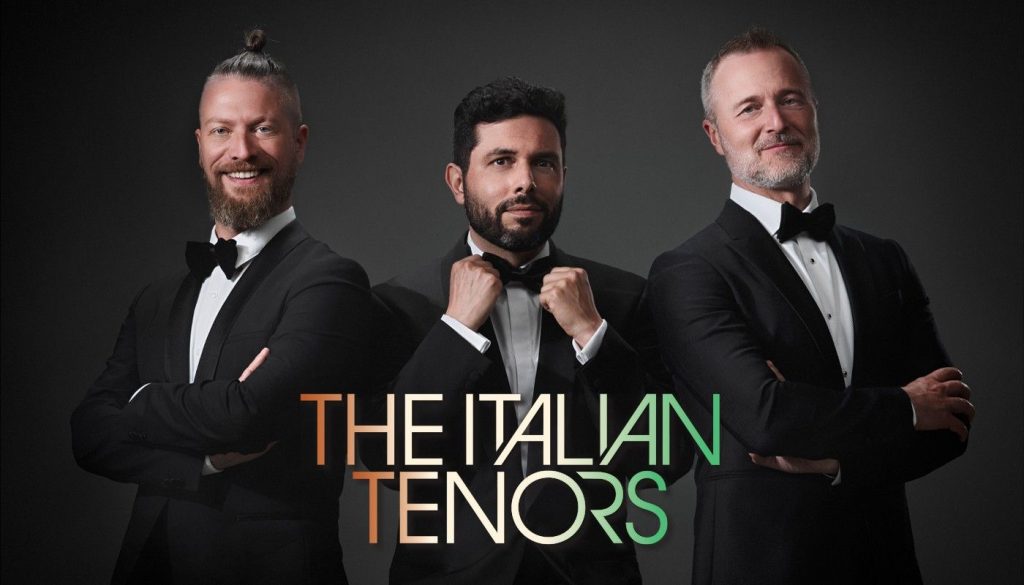 Three men in tuxedos stand against a dark background with the text "THE ITALIAN TENORS" in front of them. One adjusts his bow tie while the others stand with arms crossed.