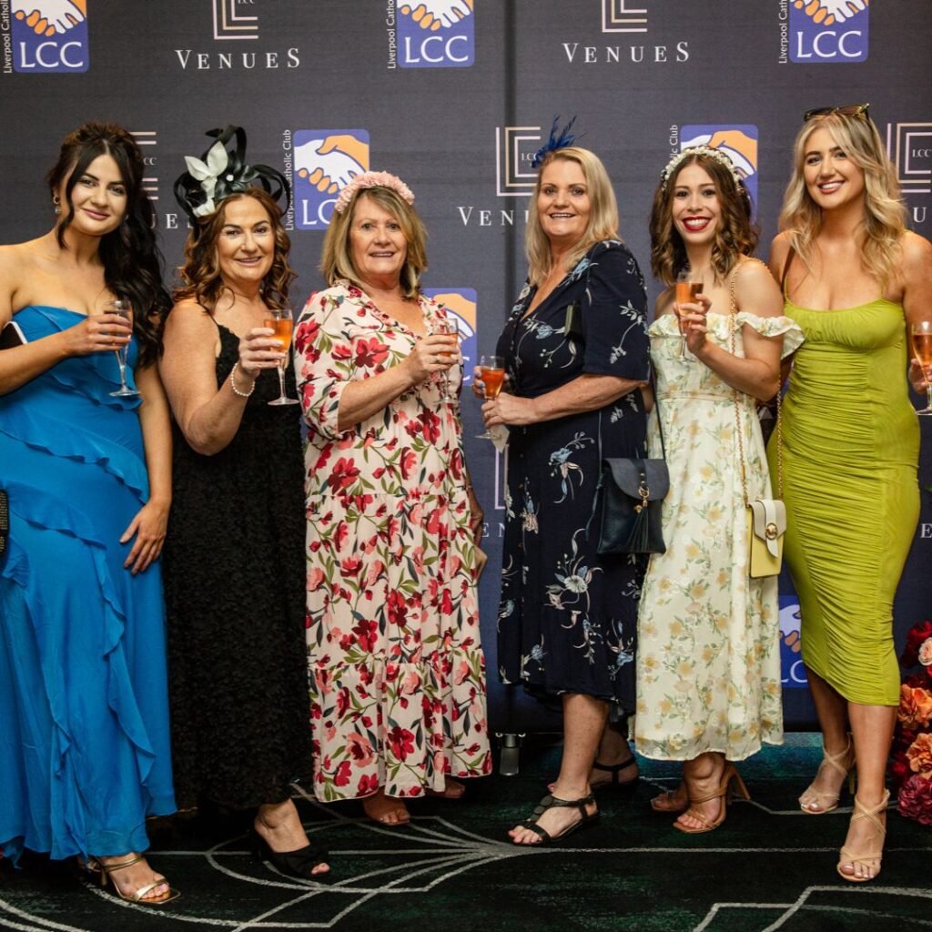 Six women in colorful dresses stand in a row, holding drinks and smiling, in front of a branded event backdrop.