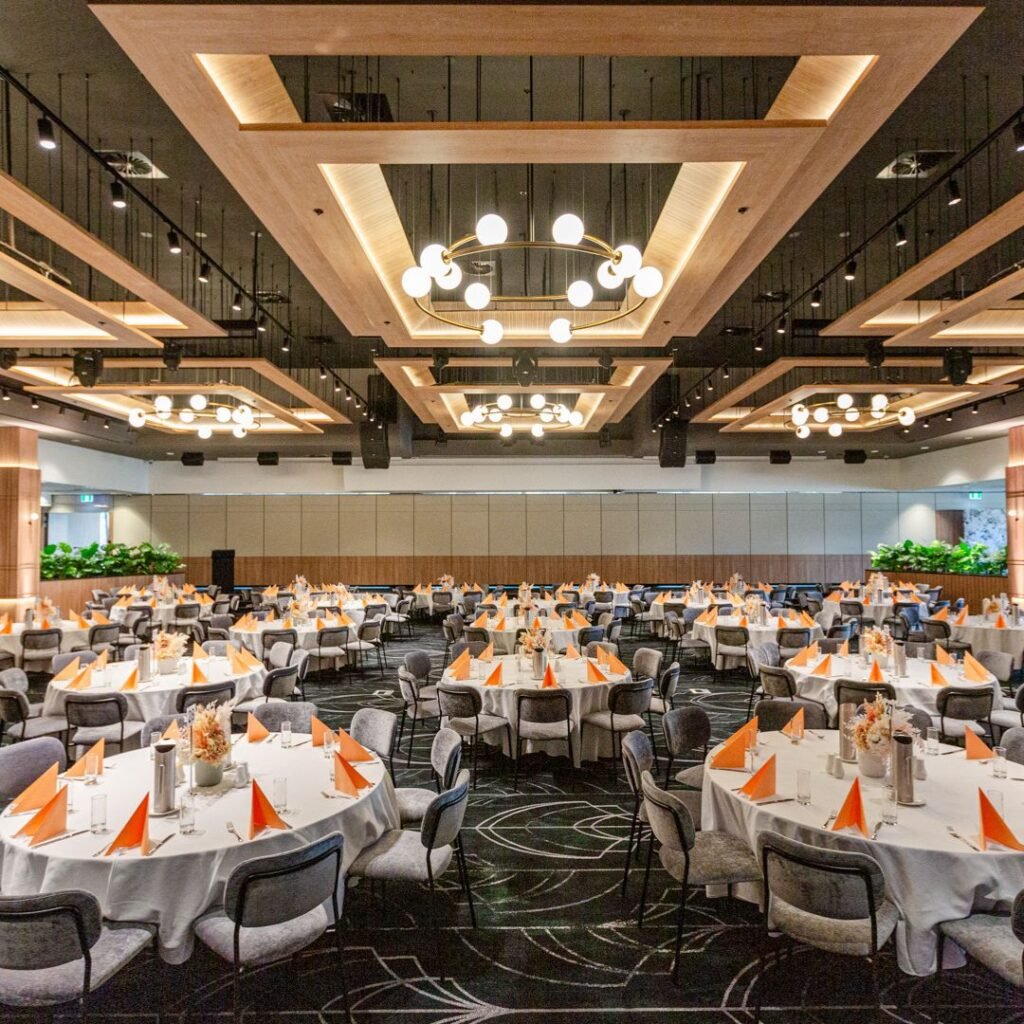 Spacious banquet hall with round tables set for an event, featuring orange napkins, modern chandeliers, and contemporary decor.