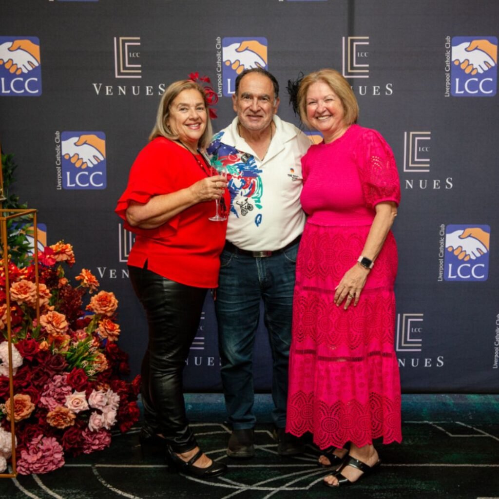 Three adults stand together and smile in front of a branded backdrop, with one woman holding a drink and flower arrangements on the left.