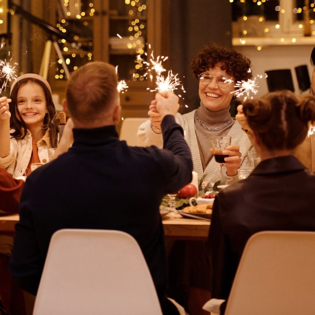A group of people sit around a dinner table holding lit sparklers and smiling, with festive lights in the background.
