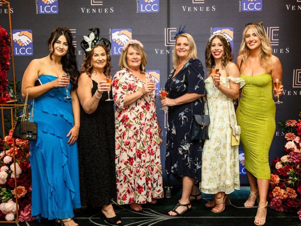 Six women in colorful dresses pose together holding drinks at an indoor event with floral decorations and branded backdrop.