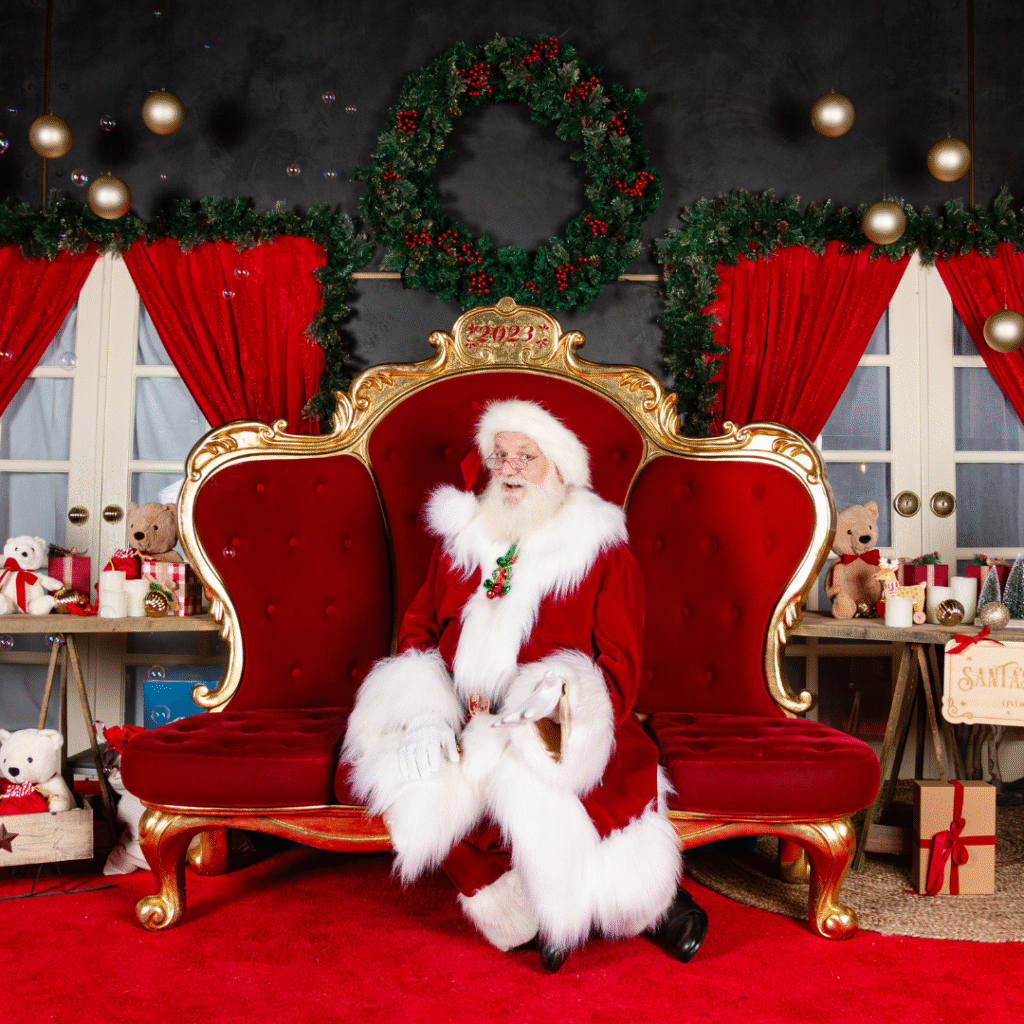 Santa Claus sits on a red and gold ornate chair in a festive room decorated with wreaths, red curtains, teddy bears, wrapped gifts, and holiday ornaments.