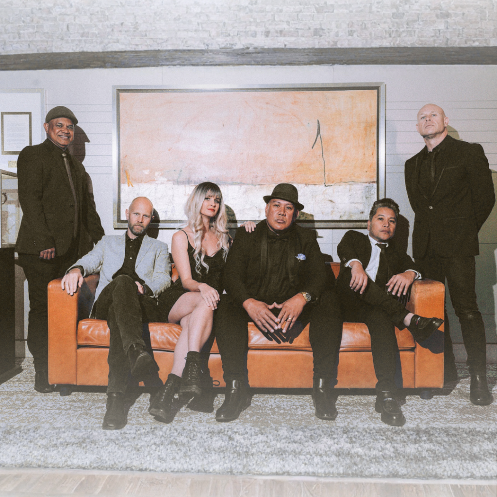 Six people in formal attire pose in a living room, with four seated on a brown sofa and two standing behind it in front of a large abstract painting on the wall.