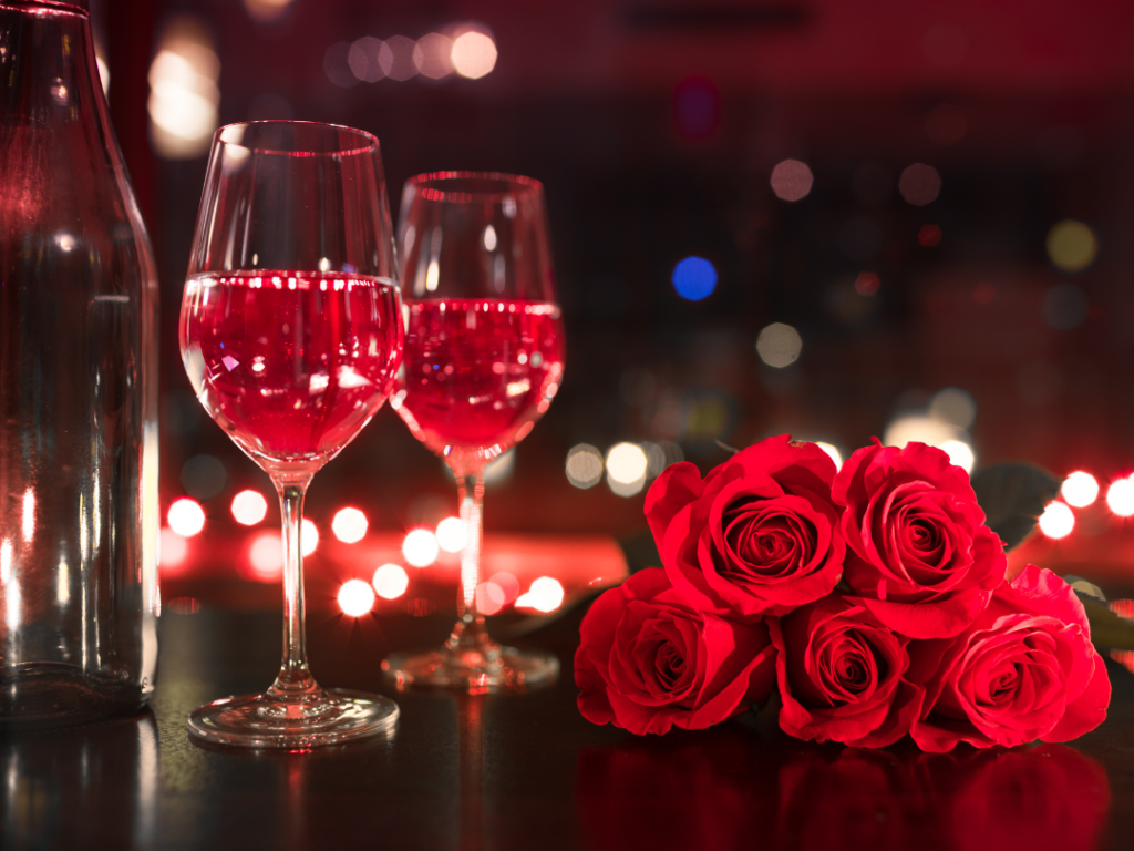 Two glasses of red wine, an empty bottle, and a bouquet of five red roses are arranged on a dark surface with blurred lights in the background.