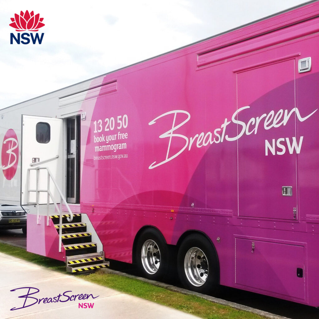 A pink BreastScreen NSW mobile clinic truck is parked with its door open and steps leading up. The BreastScreen NSW logo and contact number are visible on the side.