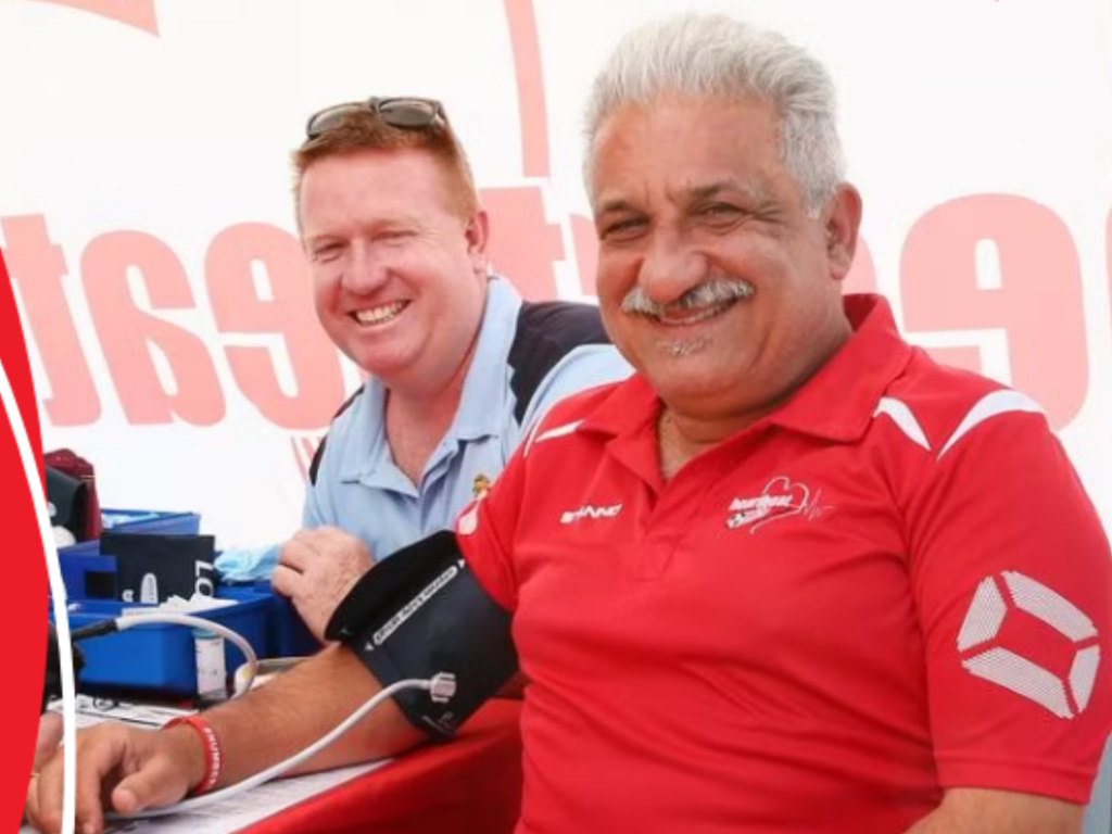 Two men sit at a table; one smiles while having his blood pressure measured, and the other sits beside him, both appearing at a public event.
