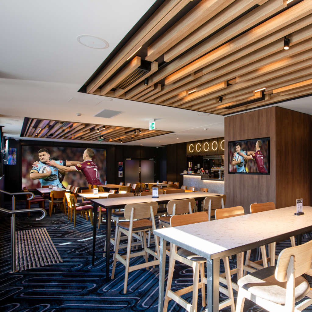 Modern sports bar interior with wooden tables and chairs, a patterned carpet, TV screens showing rugby, and a ceiling with wooden slats.