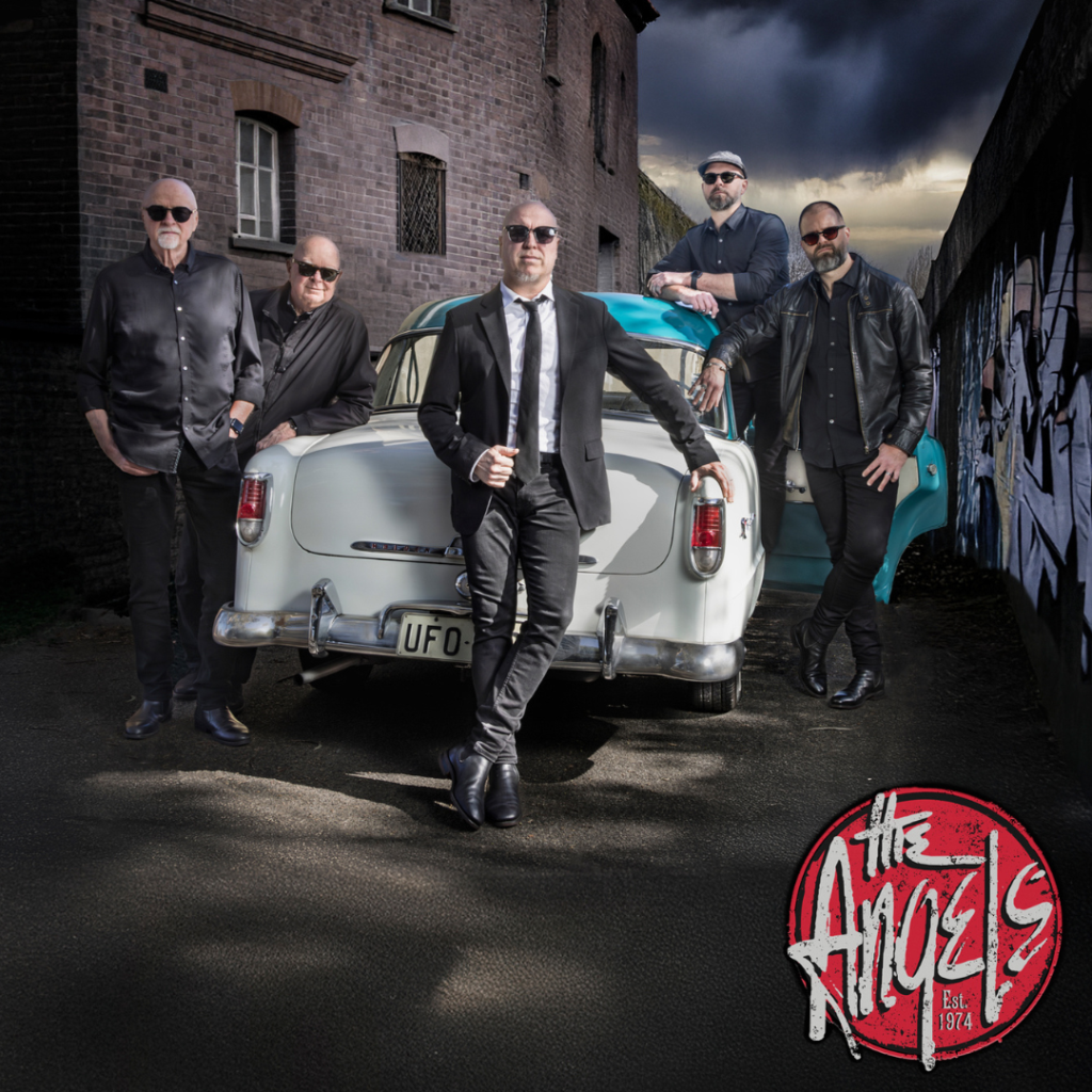 Five men in black clothing stand around a vintage white car in an alley, with "The Angels" band logo and "Est. 1974" in the lower right corner.