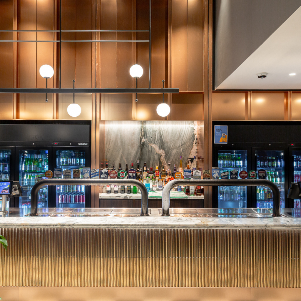 Modern bar with a marble countertop, various alcohol bottles, beer taps, and illuminated globe pendant lights, set against a backdrop of glass-fronted refrigerators.