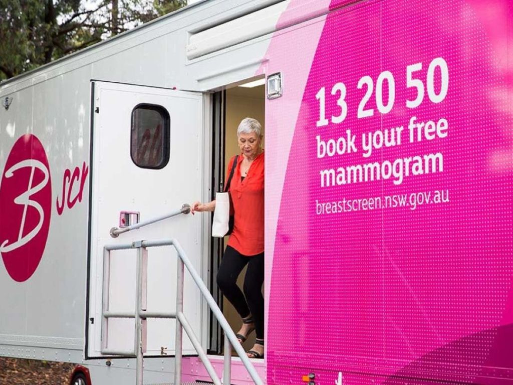 A woman exits a mobile breast screening clinic with a pink sign displaying a phone number and a website for booking a free mammogram in New South Wales.