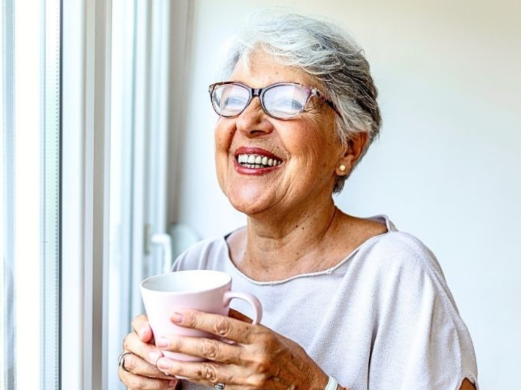 An older woman with gray hair and glasses smiles while holding a pink mug, standing by a window in daylight.