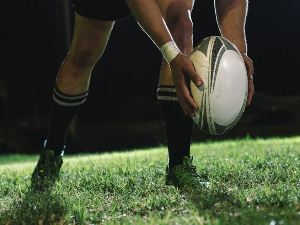 A rugby player in black shorts and striped socks bends down to place a rugby ball on the grass during a nighttime game.