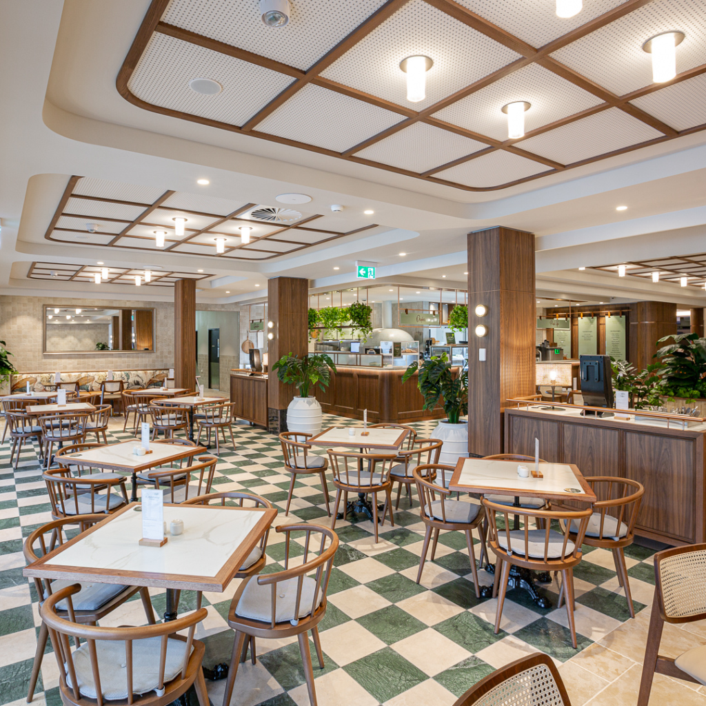 Modern, well-lit restaurant interior with wooden furniture, green and white checkered tile floor, potted plants, and an open counter area. Tables and chairs are neatly arranged.