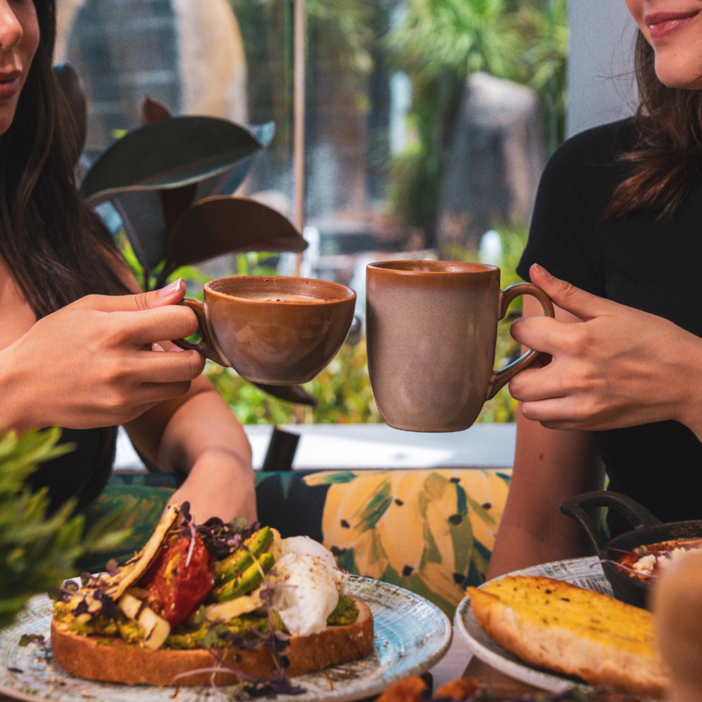 Two people sit at a table clinking ceramic coffee mugs, with plates of food including toast, avocado, and poached eggs in front of them.