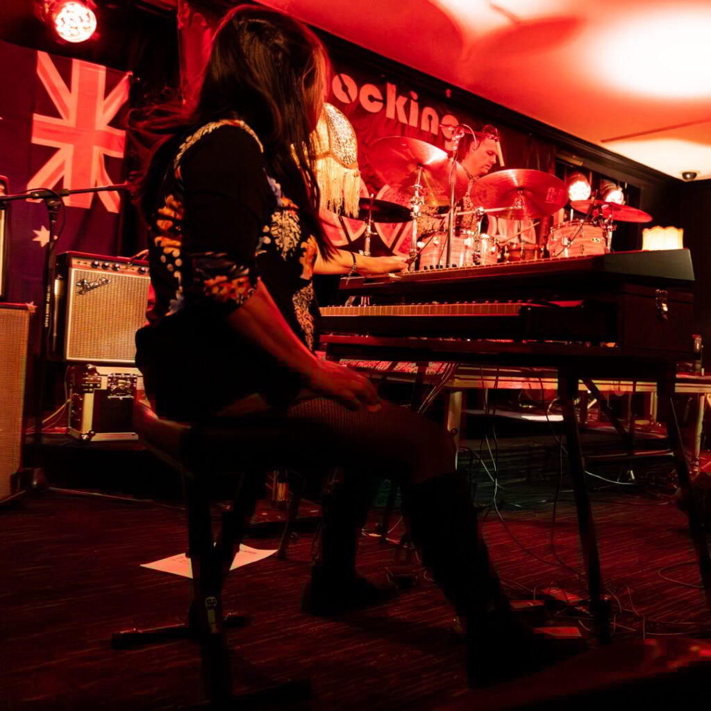 A band performs on stage under red lighting; a keyboard player sits in the foreground while the lead singer, guitarist, and drummer play in the background.