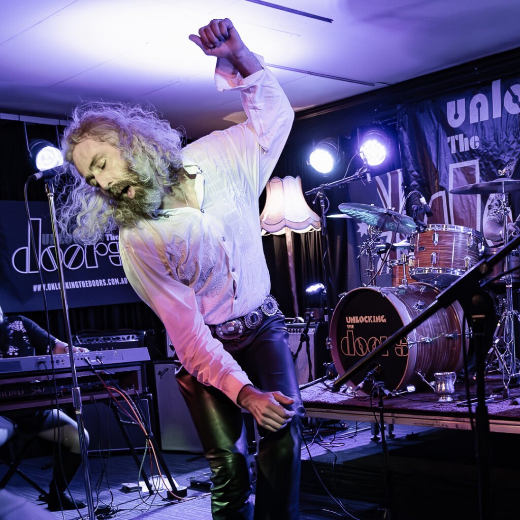 A band performs energetically on stage with a singer bending backward and a guitarist playing; audience members watch closely in a dimly lit venue.