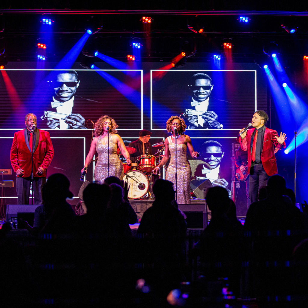 A band performs on stage with three singers in front, a drummer behind, and an image of Ray Charles displayed on screens in the background under colorful stage lights.