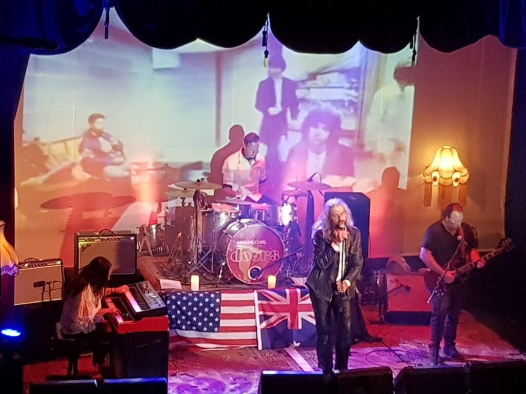 A band performs on stage with a British and an American flag draped over equipment, vintage lamps, and a projected image in the background.