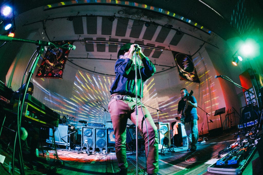A band performs on stage with colorful lights and projected patterns on the wall behind them; one person sings into a microphone at the center.
