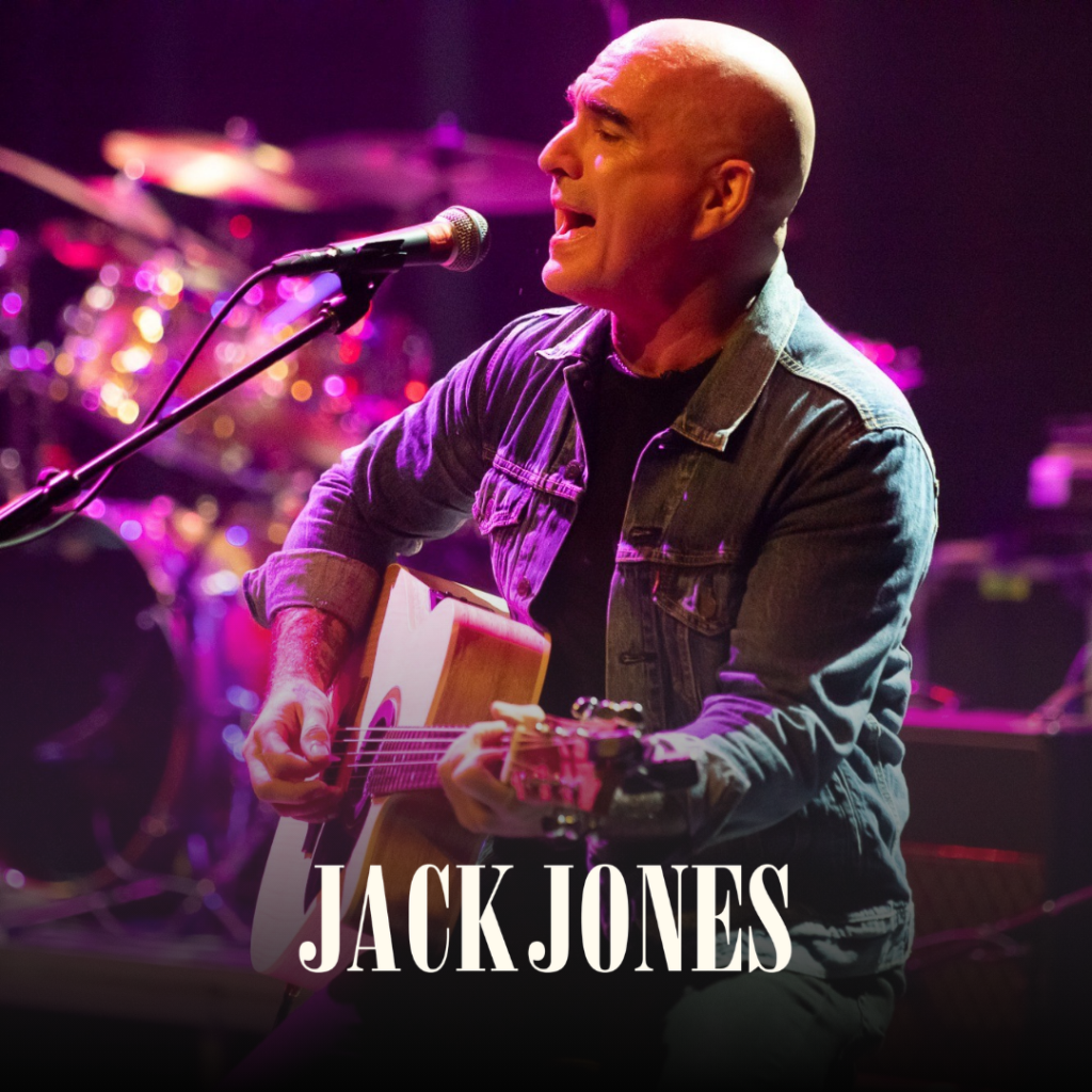 A man plays an acoustic guitar and sings into a microphone on stage. He wears a denim jacket. The name "Jack Jones" is written in large white text at the bottom.