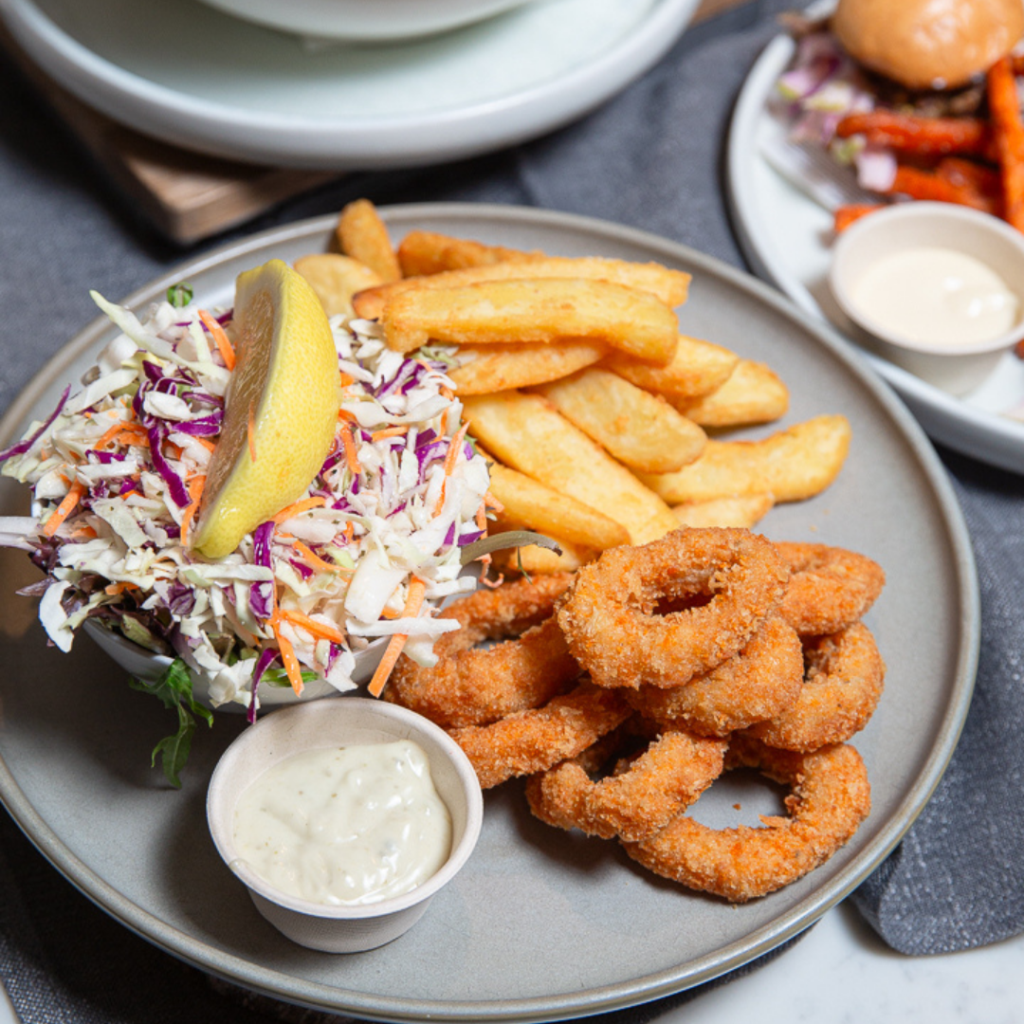 A plate with fried onion rings, thick-cut fries, coleslaw topped with a lemon wedge, and a small cup of dipping sauce.
