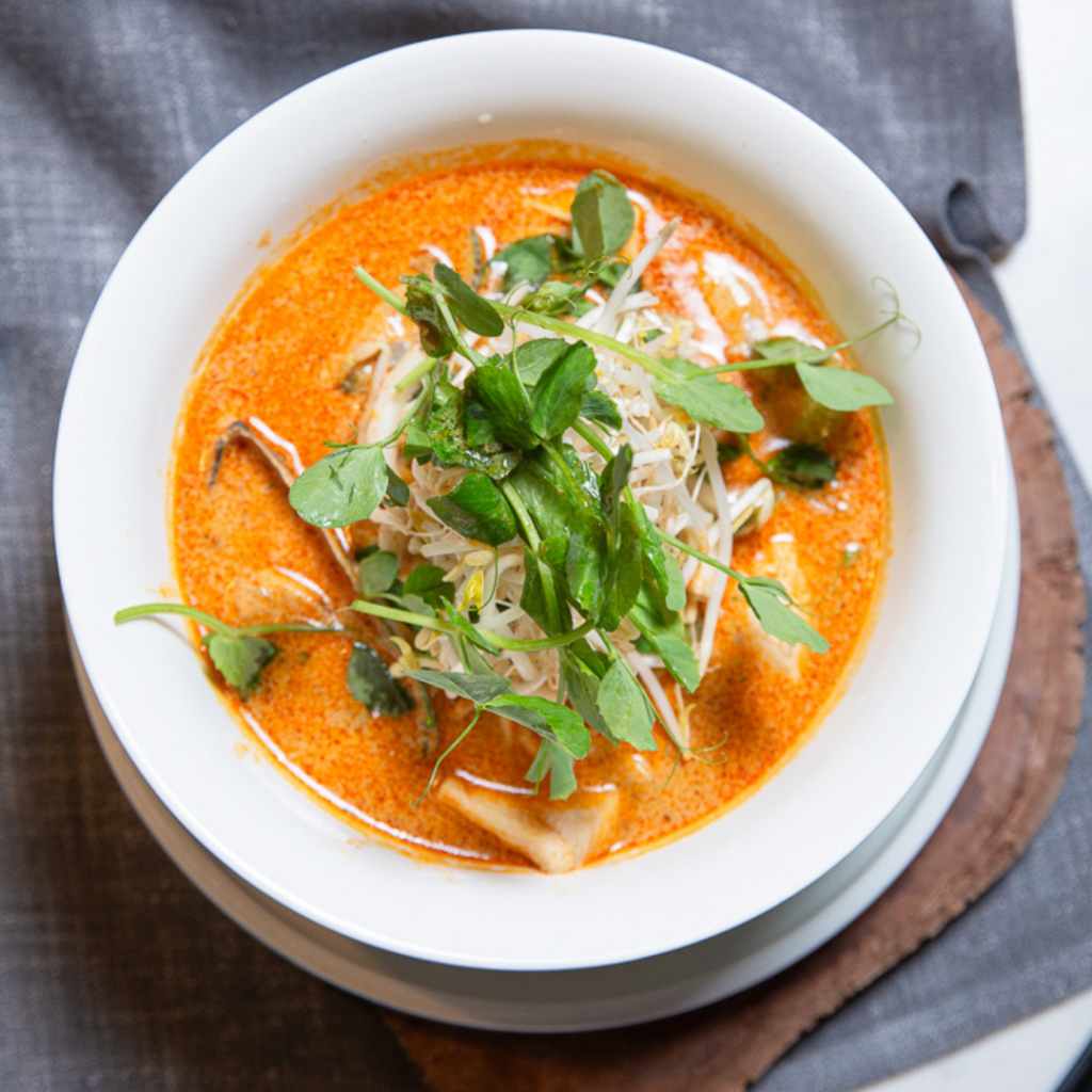 A bowl of creamy orange soup topped with herbs and sprouts, placed on a gray napkin and round wooden board.