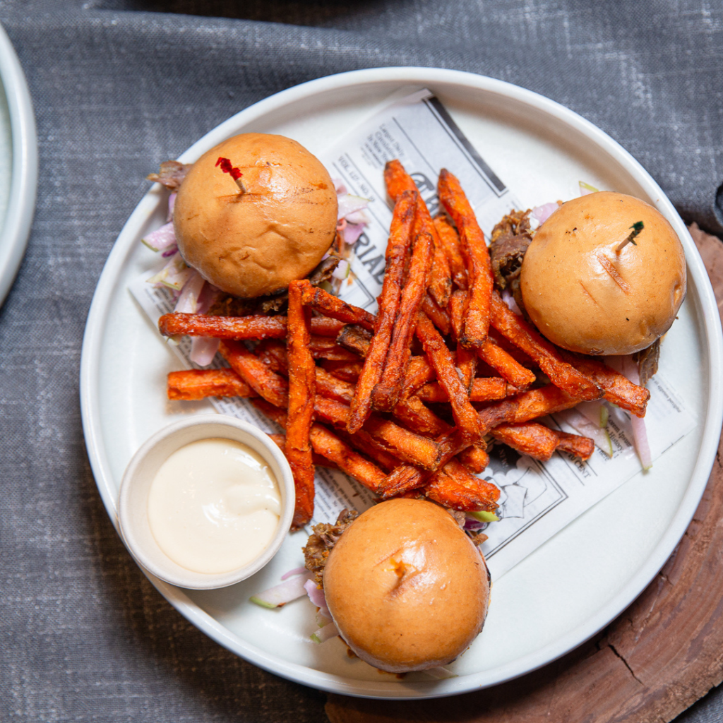 Three mini burgers with shredded lettuce, sweet potato fries, and a side of dipping sauce arranged on a white plate lined with newspaper-print paper.