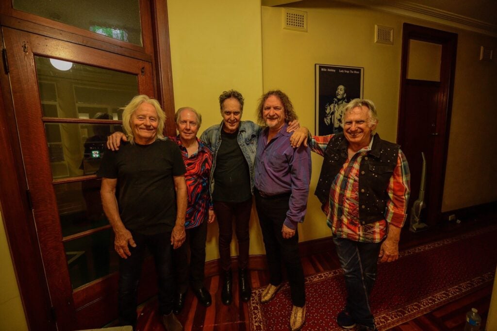 Five older men standing together and smiling in a warmly lit hallway, with a framed poster on the wall