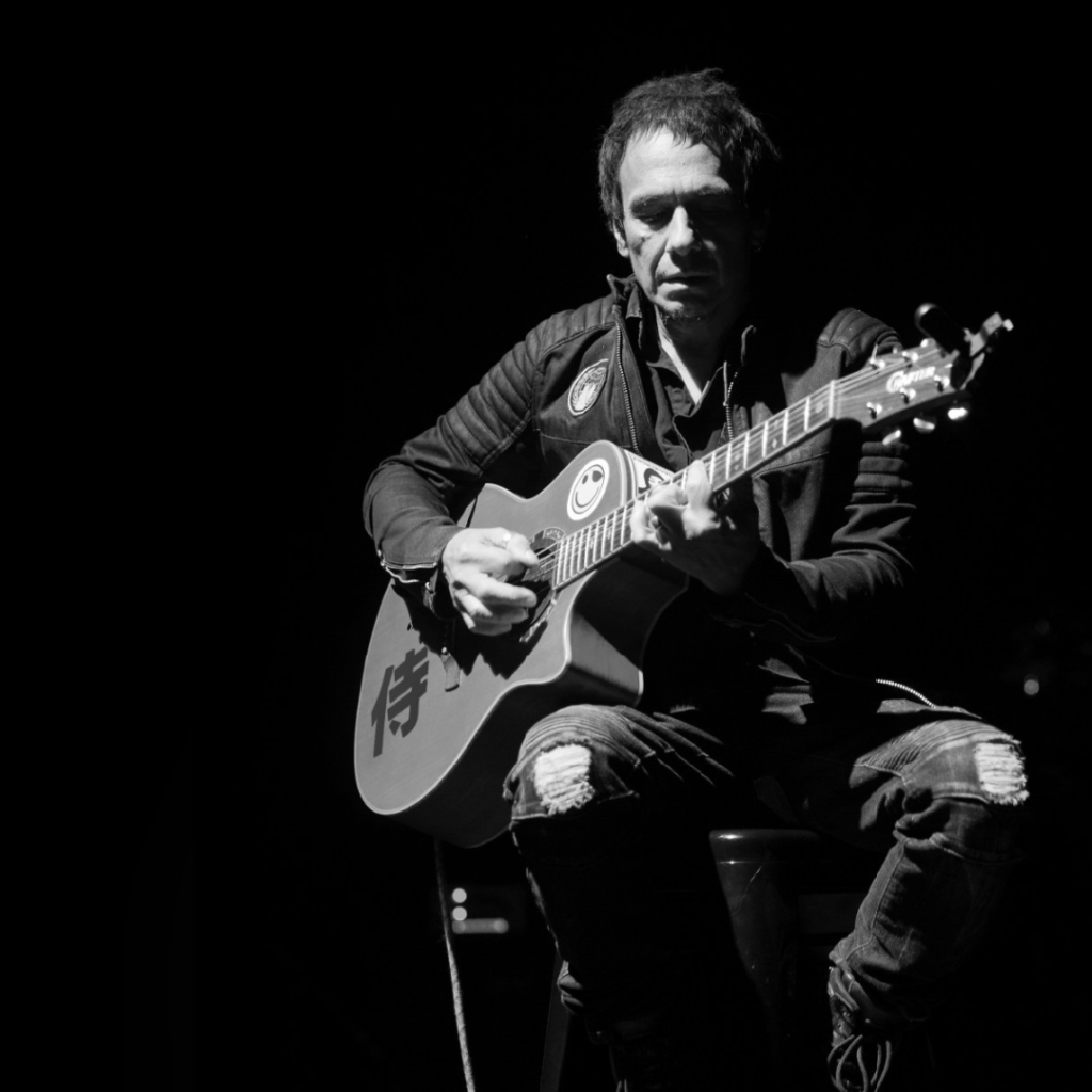 A person sits on a stool playing an acoustic guitar. The image is in black and white, and the scene is dimly lit with the background mostly dark.