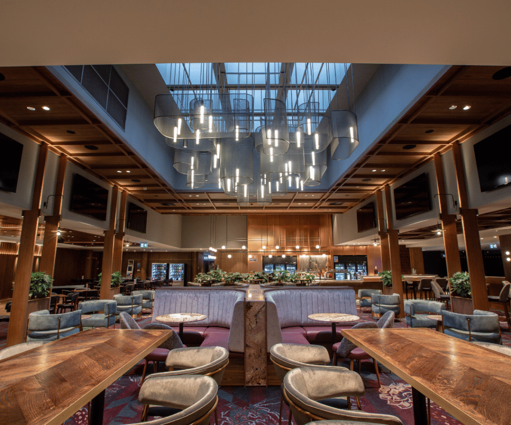Modern restaurant interior with wooden tables, upholstered seating, pendant lights, and a skylight ceiling, featuring a symmetrical, open design and decorative plants.