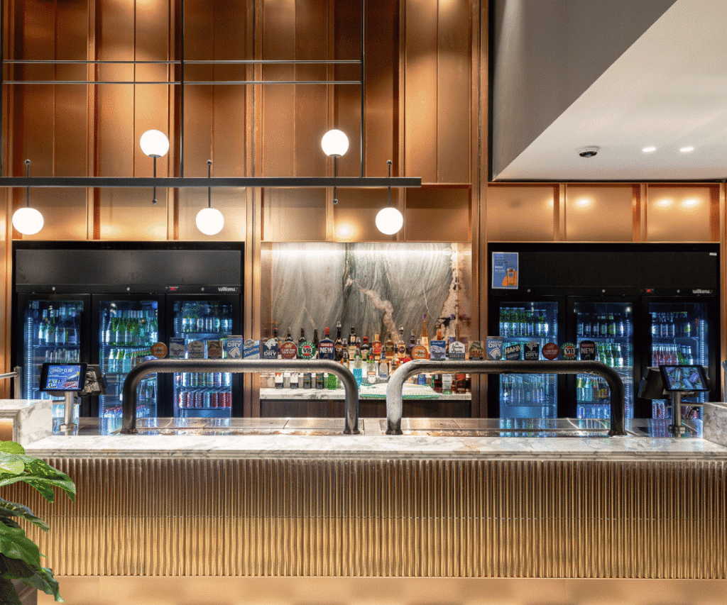 Modern bar with a marble countertop, beer taps, and illuminated shelves stocked with bottles and cans. Hanging globe lights and metallic accents decorate the space.