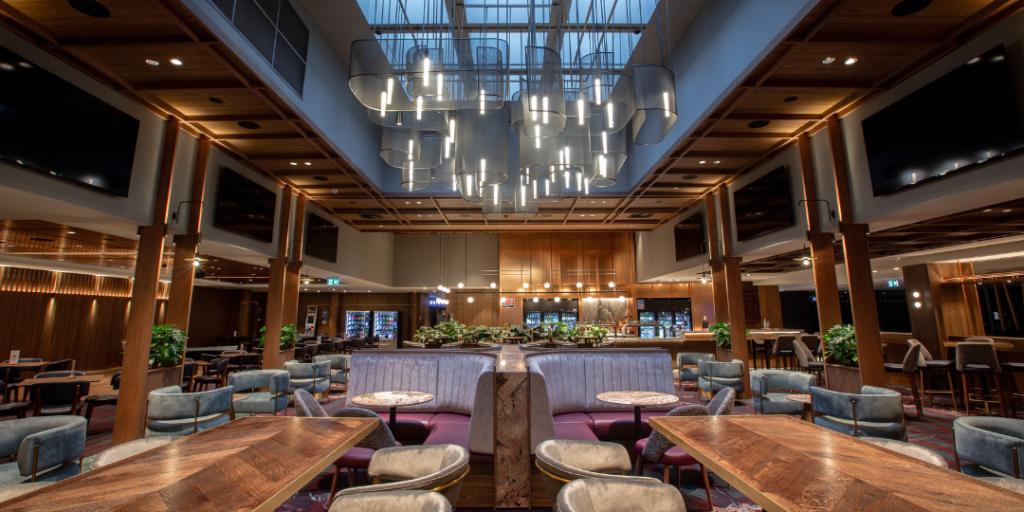 Spacious modern restaurant with wooden tables, upholstered booths, plush chairs, large hanging lights, and a high ceiling with skylights.