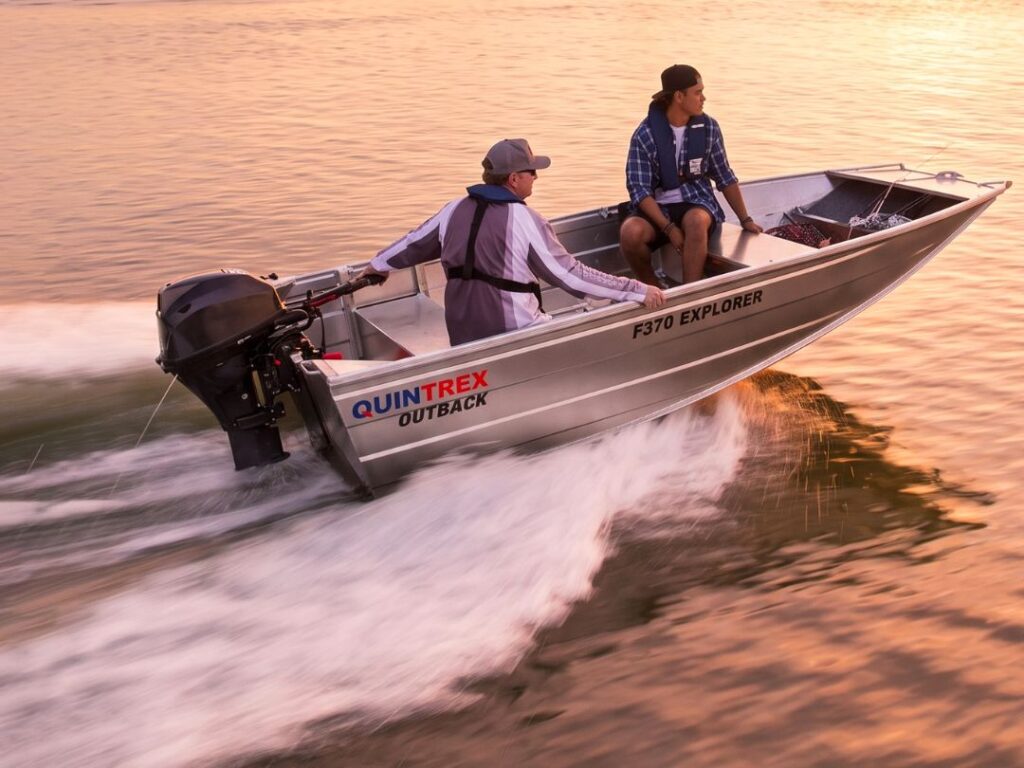 Two people ride a small aluminum motorboat labeled “QUINTREX OUTBACK F370 EXPLORER” on calm water at sunset, creating a wake behind them.