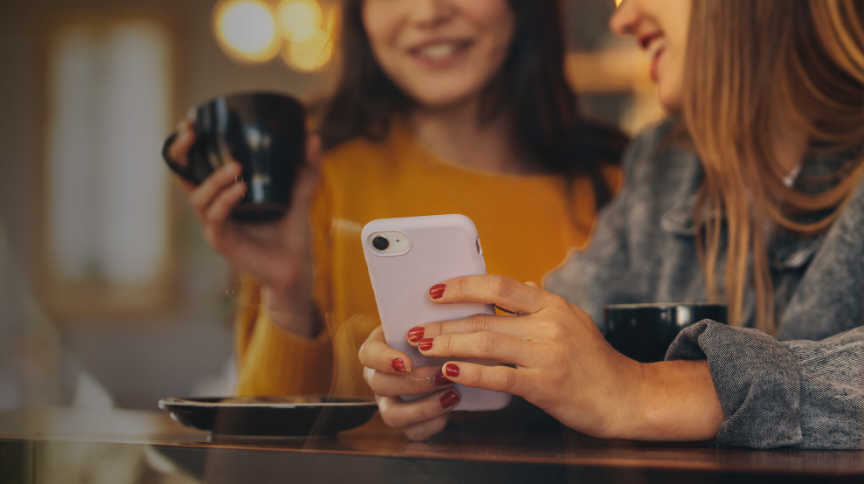 Two women sitting at a table, holding coffee mugs and looking at a smartphone together, smiling.