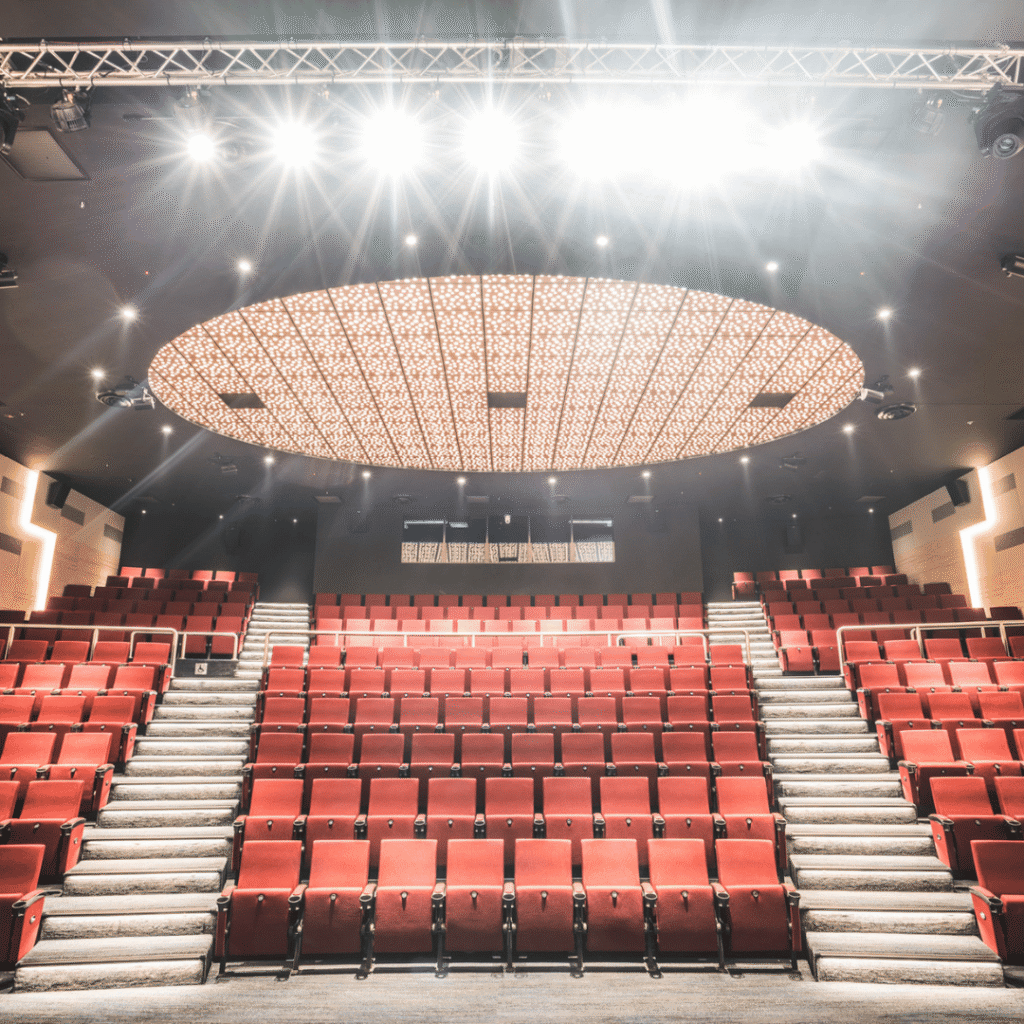 Empty theater with rows of red seats, modern lighting above, and an illuminated oval ceiling feature. Photo is taken from the stage facing the audience seating.